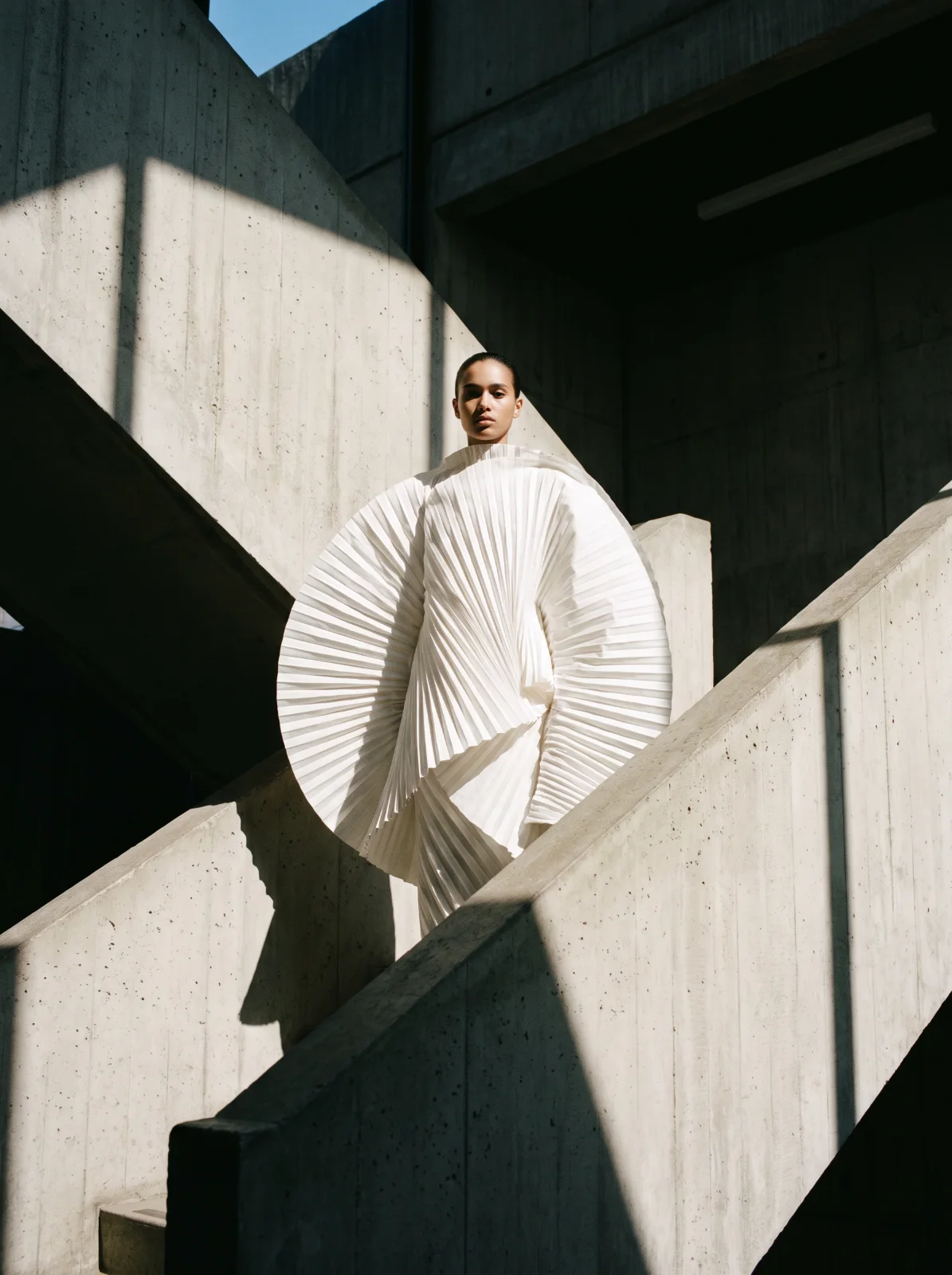 Fashion editorial: a sculptural white pleated garment in a sunlit concrete stairwell.